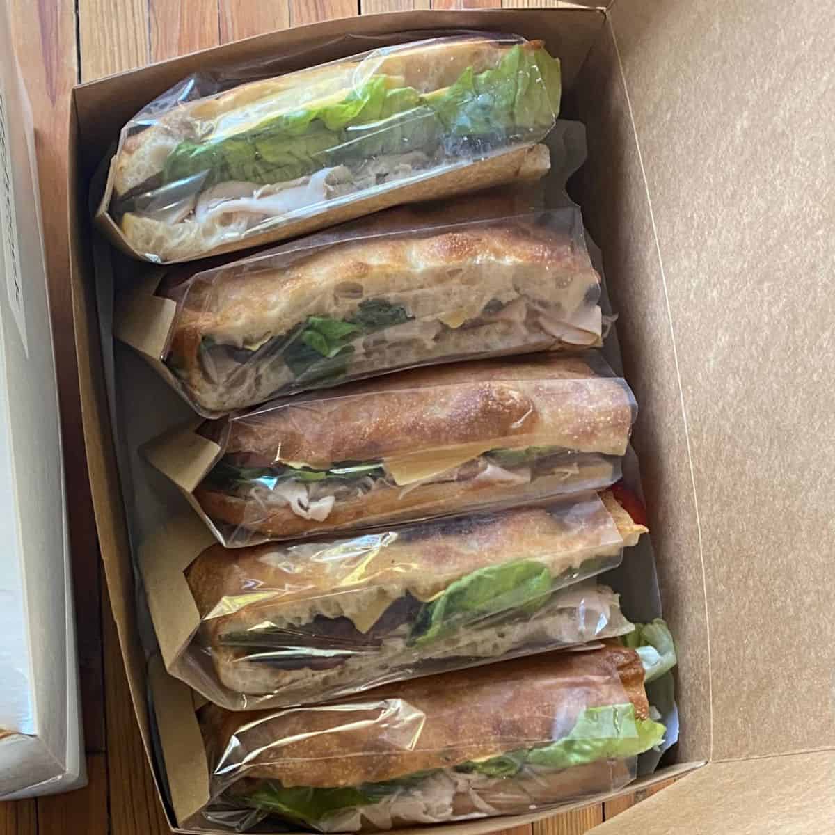 Six turkey and cheese focaccia sandwiches with lettuce and tomato, packaged in a box.