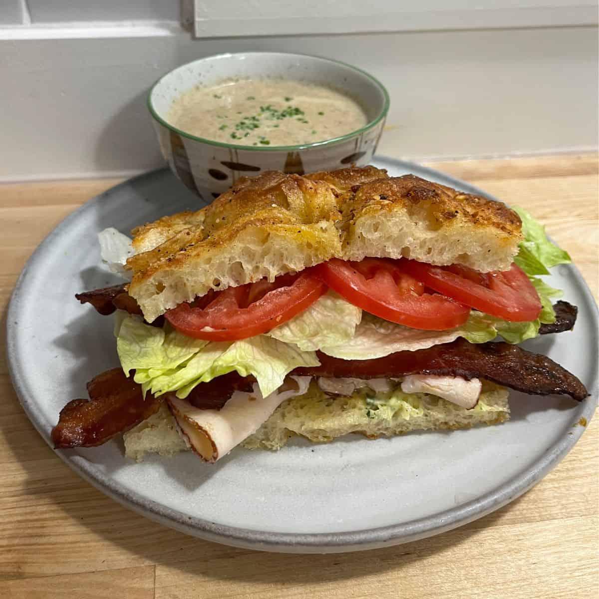 A turkey, bacon, lettuce and tomato on focaccia with a cup of soup in the background.