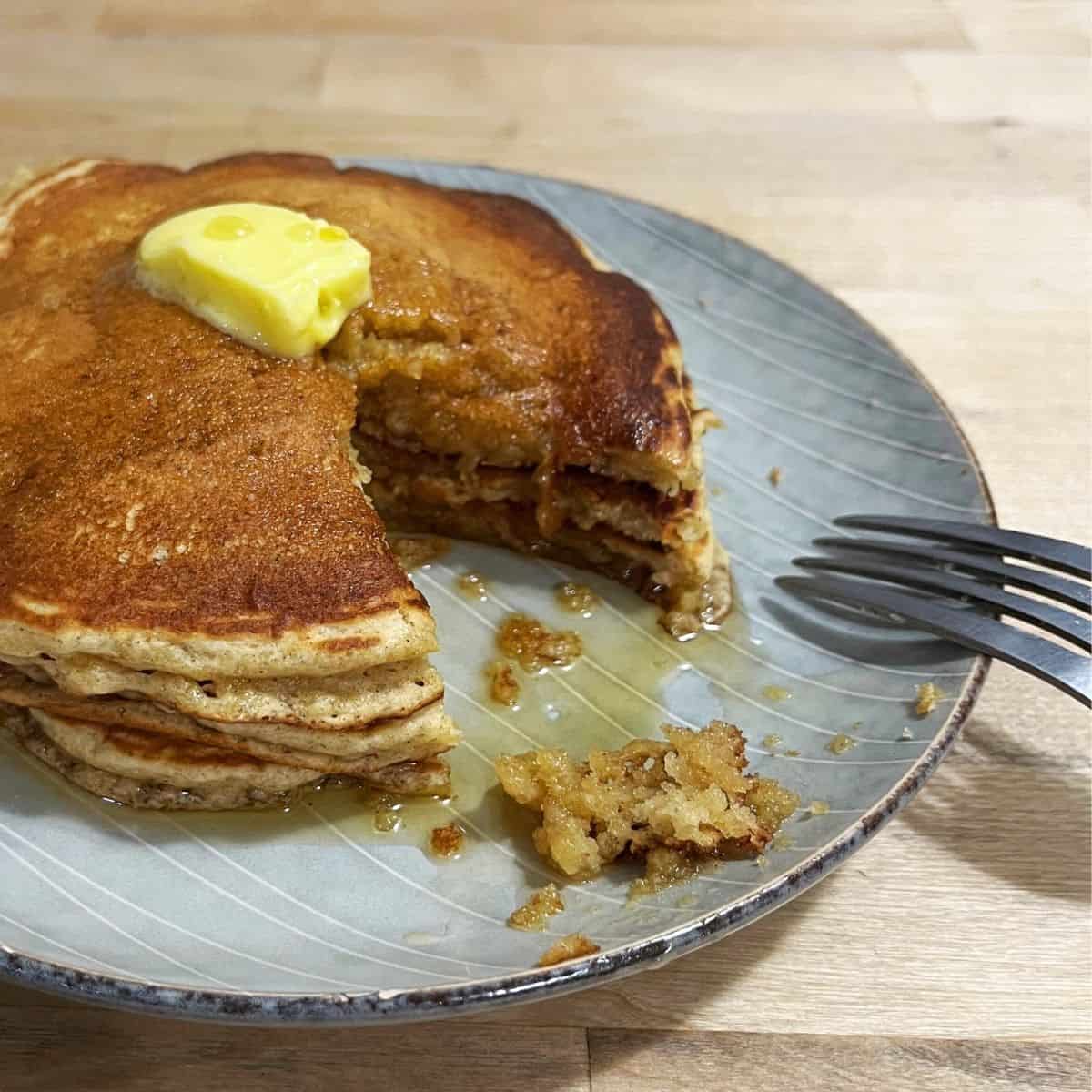 A stack of three whole wheat banana pancakes with a pat of butter and maple syrup.  The pancakes have been cut into, showing the interior.  There's a fork on the plate.
