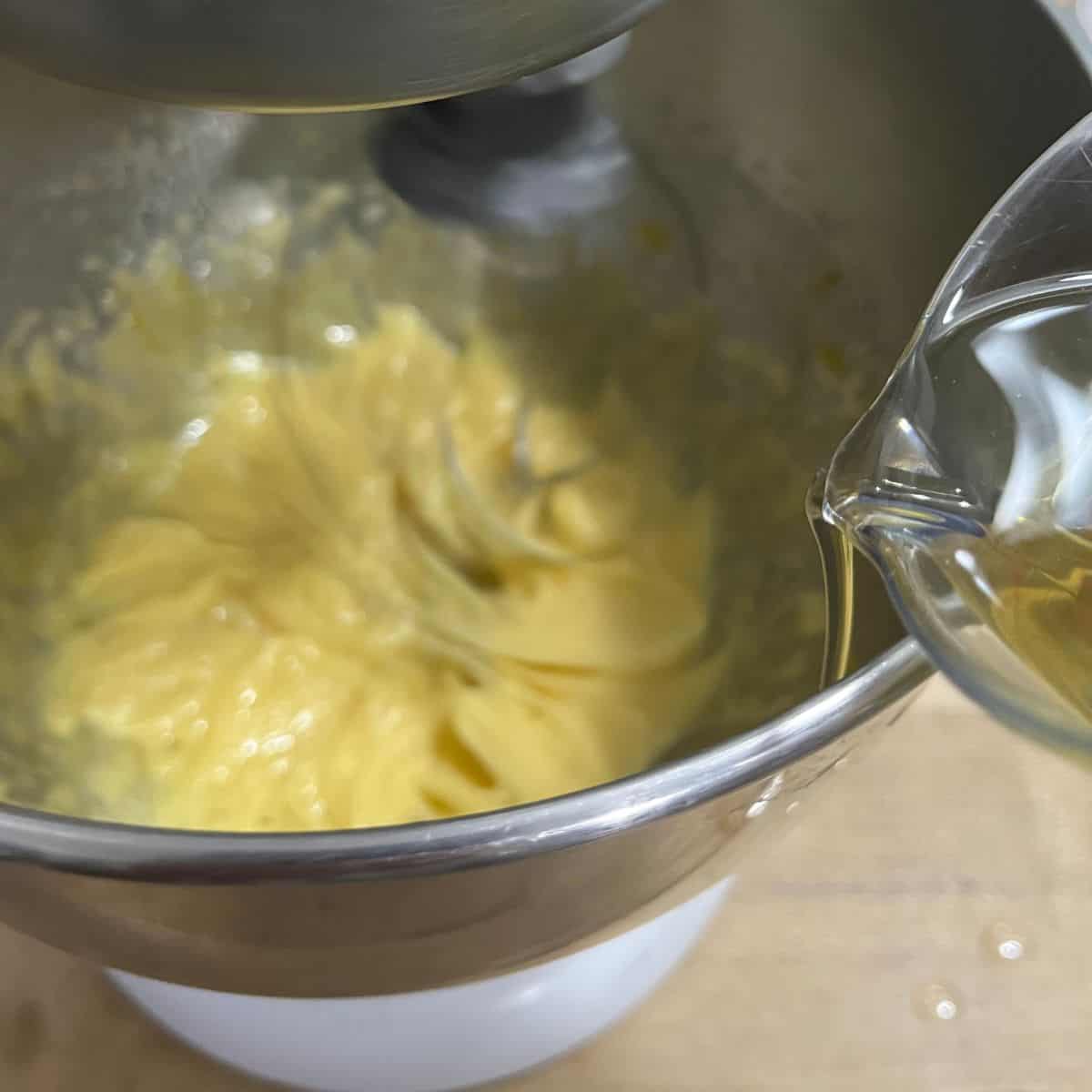 The yolk mixture is a paler yellow.  The whip is still mixing and oil is being drizzled in on the side of the bowl.