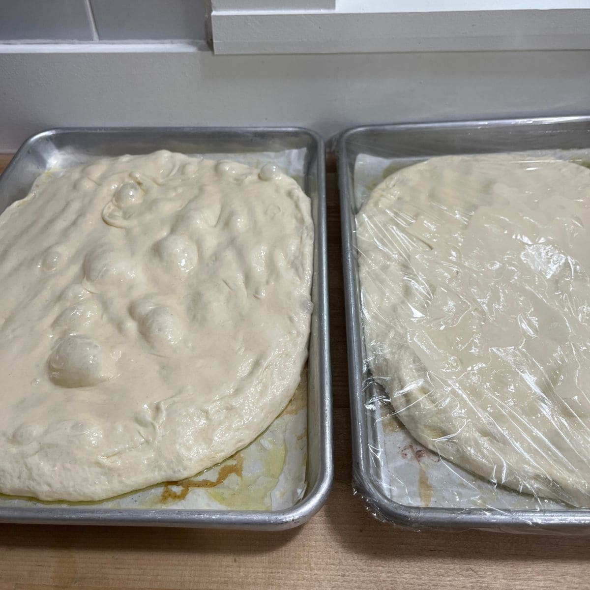 Two trays of focaccia dough, flattened and stretched ¾ of the way.  One dough shows plastic covering it.
