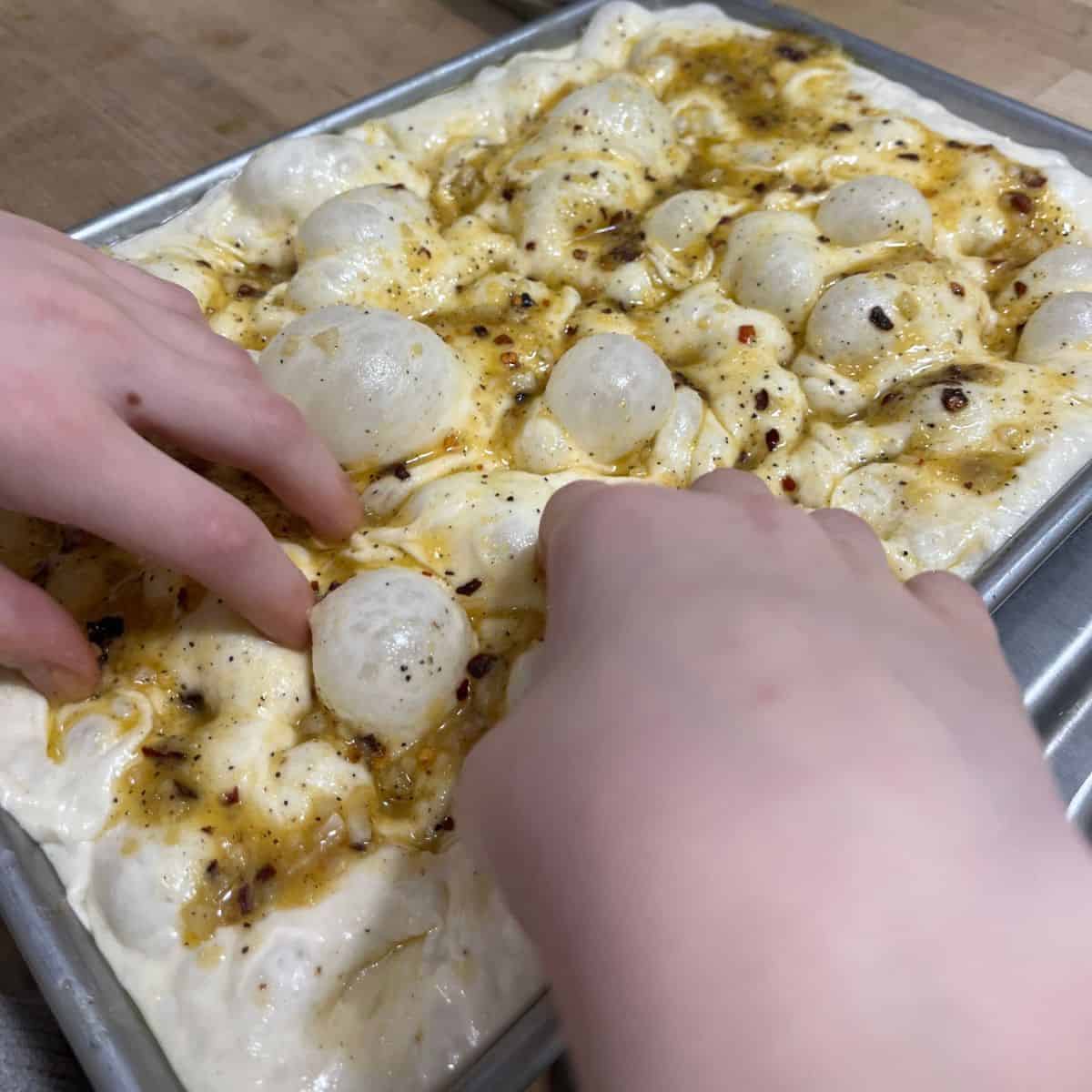 Two hands dimpling focaccia dough.  Dough fills the pan completely and is showing bubbles.