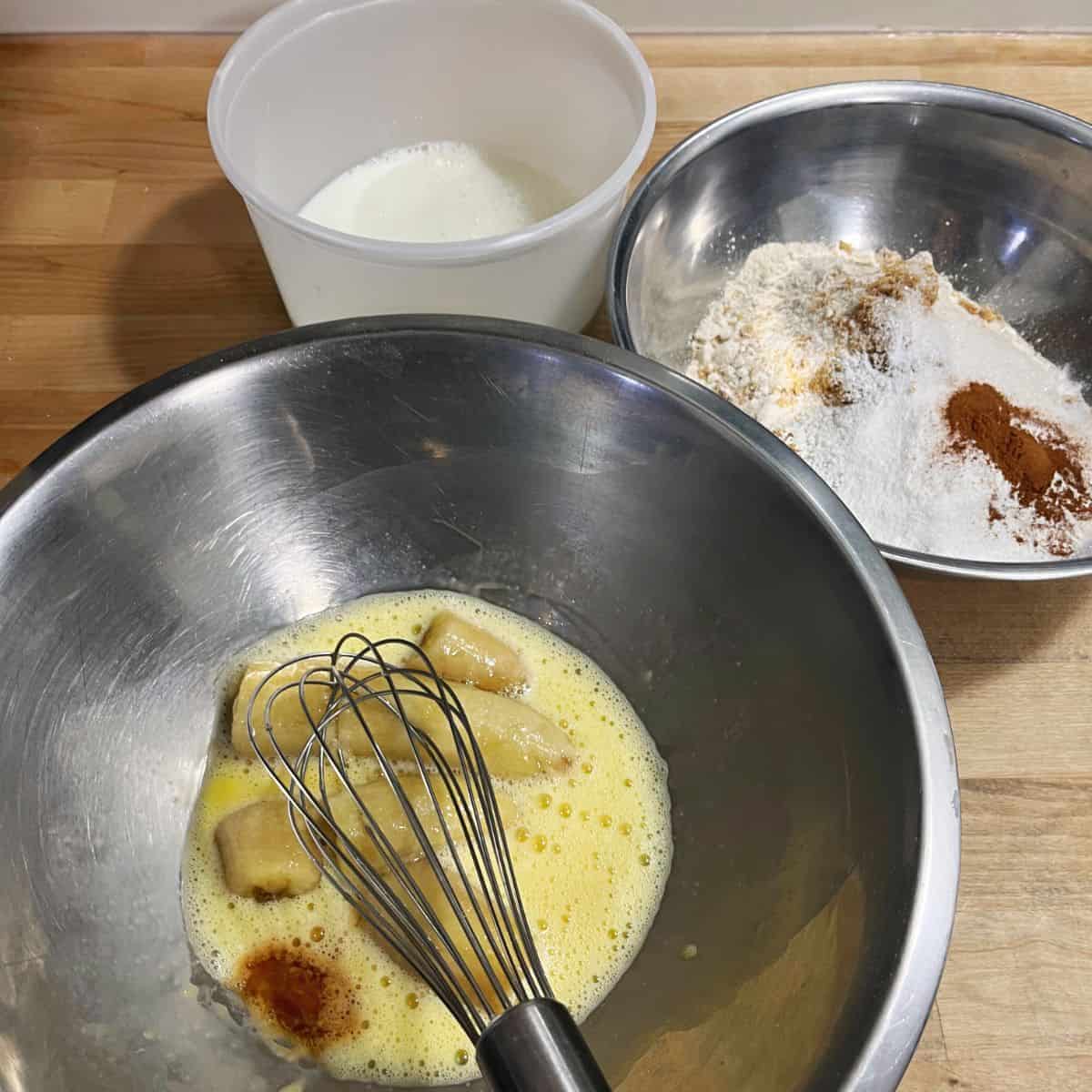 A bowl with a whisked egg, banana, vanilla and melted butter. A second bowl with whole wheat flour, brown sugar, cinnamon, baking power, baking soda and salt. A third container with milk + vinegar.