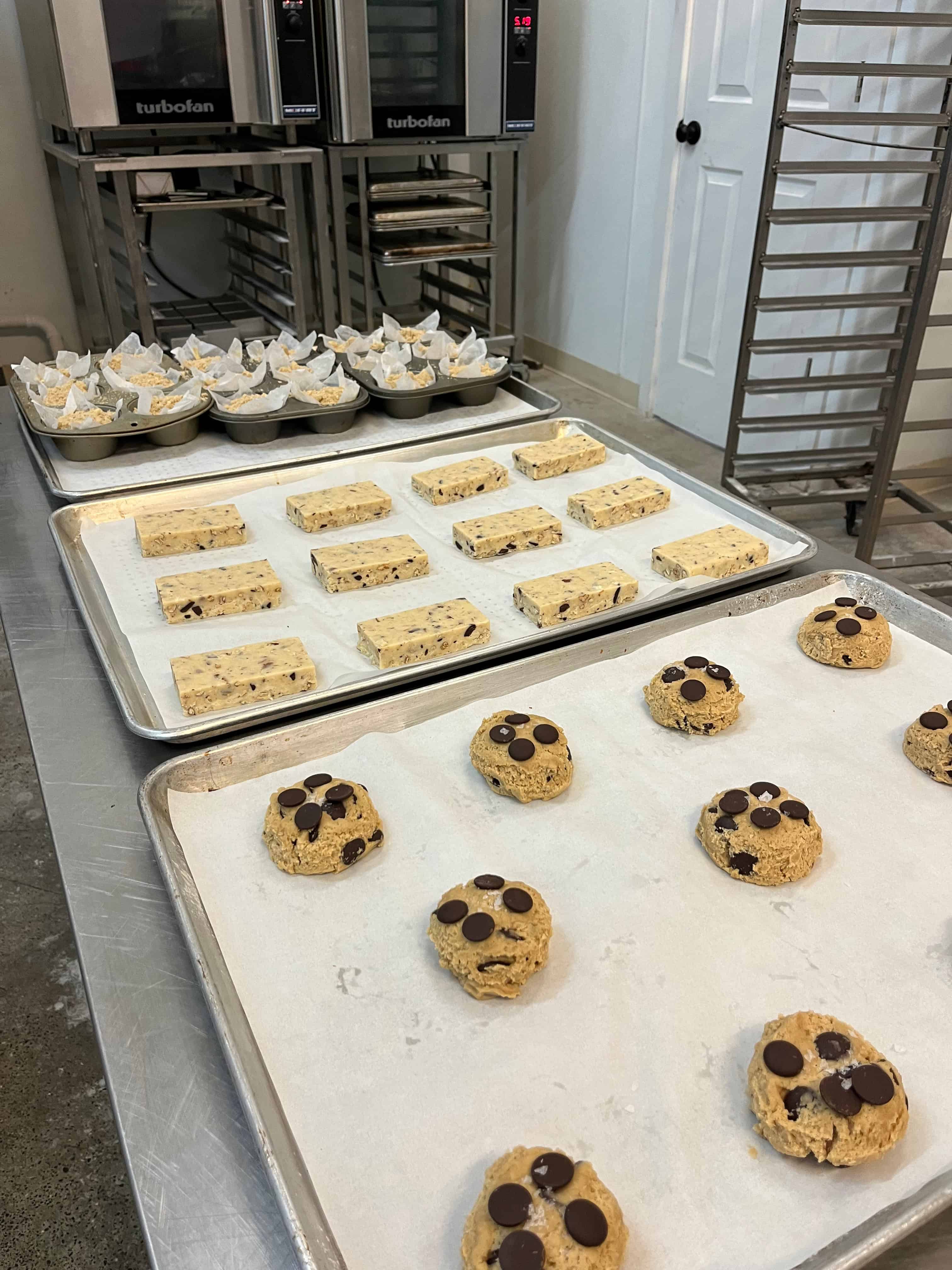 One tray of chocolate chip cookies, one tray of shortbread, one tray of coffee cake muffins with ovens and baking rack in background