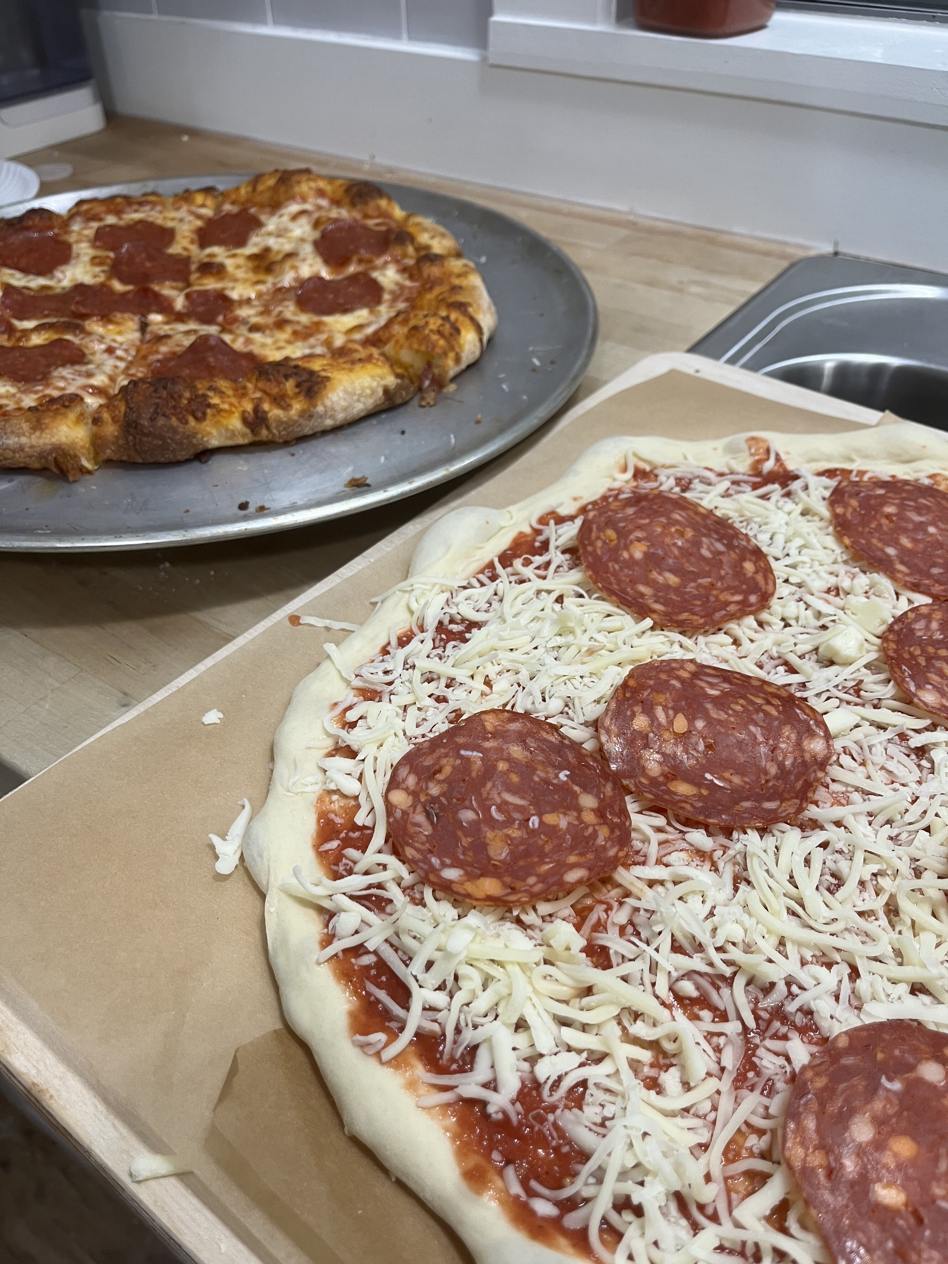 Assembled, raw pizza in the foreground and a baked pizza in the background.